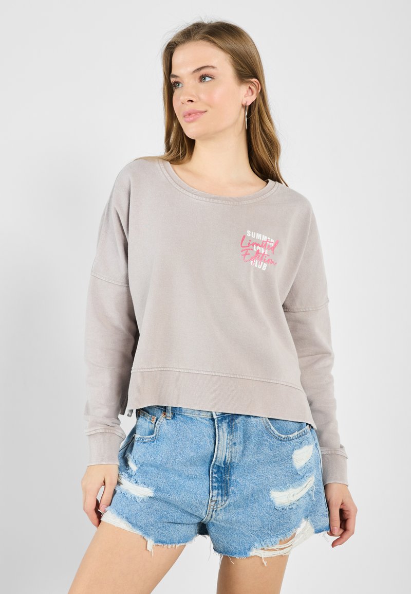 Lys grå cropped sweatshirt med lange ermer og rund hals, med en rosa grafisk trykk på brystet. Brukt over slitte denimshorts.