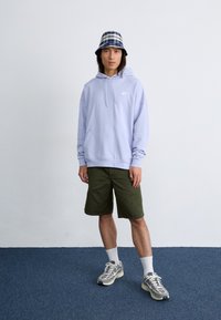 Young man stands against a plain wall, wearing a light purple hoodie, green shorts, and a plaid bucket hat, with sneakers and white socks.