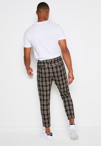 Man som bär en vit t-shirt, svart och vit rutiga byxor samt vita Reebok-sneakers, stående mot en neutral vit vägg, med ryggen vänd mot betraktaren.