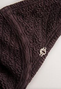 Tissu tricoté brun foncé texturé avec une bordure côtelée et un petit pendentif rond en métal argenté fixé près du bord.