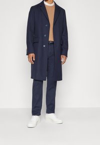 Marinblå ullkappa med slagkrage och knappar, i kombination med en beige tröja, marinblå byxor och vita sneakers. Ren siluett och skräddarsydd passform.