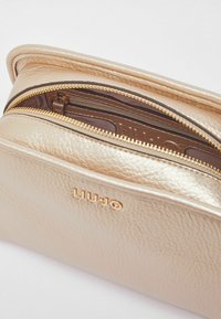 Bolso de cuero tono dorado con acabado texturizado, con cierre de cremallera y logo de la marca grabado en el frente.