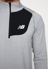 Pullover cinza com fecho, com um padrão texturizado e um painel preto no ombro esquerdo. Apresenta um logo branco "NB" na área do peito.