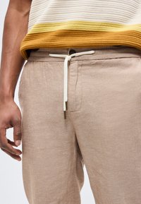 Uomo con pantaloni beige con coulisse e maglia a righe marrone e bianca, mano rilassata lungo il fianco.