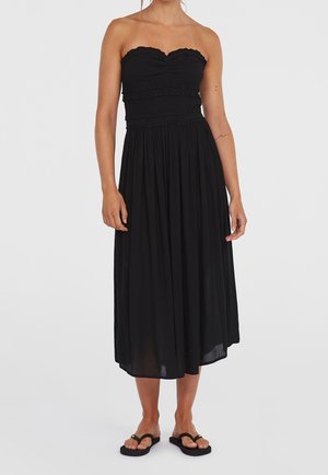 Sort stropløs midi-kjole med rynket overdel og samlet nederdel, båret med sorte flade sandaler på en stående person med bare skuldre.