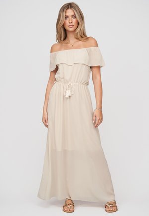 Femme blonde portant une robe maxi beige épaules dénudées avec ceinture à pompons et sandales dorées, debout devant un fond clair uni.