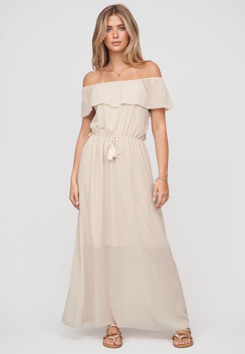 Femme blonde portant une robe maxi beige épaules dénudées avec ceinture à pompons et sandales dorées, debout devant un fond clair uni.