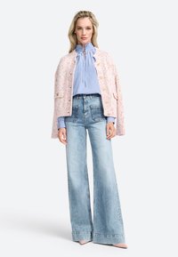 Gilet en tricot rose avec des éclats multicolores, blouse rayée bleue avec un col noué, et jeans en denim clair à jambes évasées avec de grandes poches.