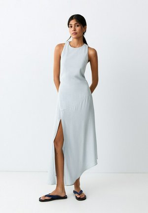 Kvinde med langt mørkt hår i en lyseblå ærmeløs kjole med sidelommer, stående barfodet på en hvid gulv mod en hvid væg.