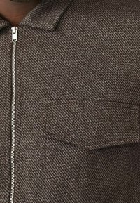 Donkerbruin shirt met rits en een gestructureerd visgraatpatroon, voorzien van een kraag en een borstzak. Zilverkleurige ritssluiting als accenten.