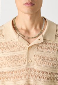 Beige knitted polo shirt with decorative lace pattern, featuring two buttons and a fold-down collar. Layered necklace with chain and pearls.