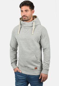 Jasnoszara bluza z kapturem, kremowymi sznurkami, kieszenią z przodu oraz brązową skórzaną metką na dole. Materiał z mieszanki bawełny.
