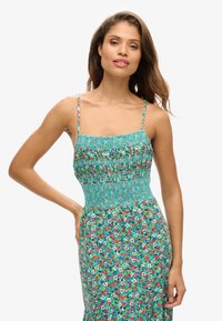 Kwiatowa sukienka maxi z turkusowym marszczonym gorsetem, cienkimi ramiączkami oraz żywym wzorem kwiatowym w kolorze czerwonym, różowym i białym.
