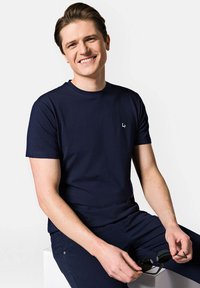 Granatowa koszulka z krótkim rękawem wykonana z bawełny, z małym białym logo na piersi. Model trzyma okulary przeciwsłoneczne, siedząc na białej powierzchni.