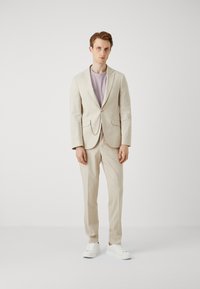Beige linnen pak met lichtpaarse T-shirt, voorzien van een enkele knoopsluiting en zijzakken; gecombineerd met witte sneakers.