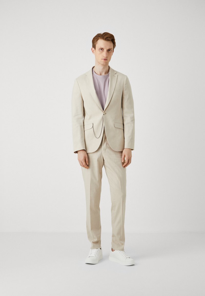 Beige linnen pak met lichtpaarse T-shirt, voorzien van een enkele knoopsluiting en zijzakken; gecombineerd met witte sneakers.