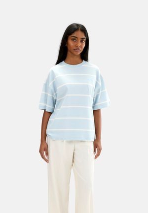 Kvinde iført en lyseblå oversized T-shirt med hvide vandrette striber og løstsiddende cremefarvede bukser, stående mod en ensfarvet baggrund.