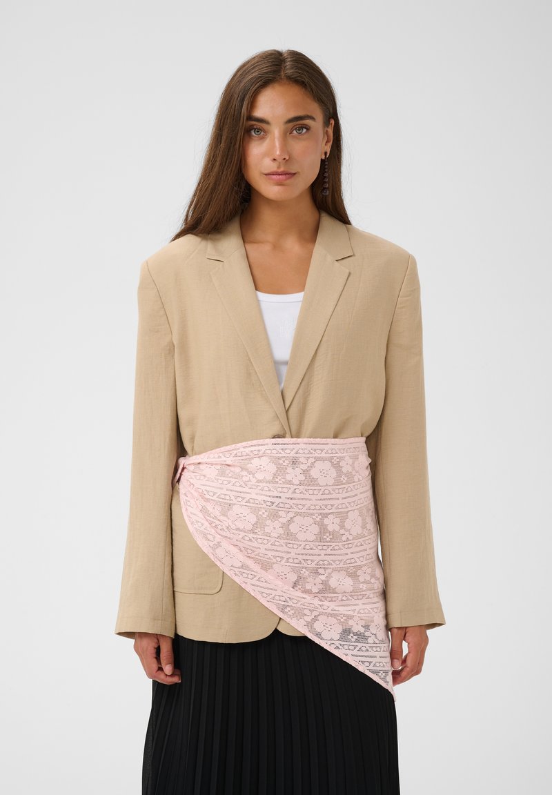 Femme aux longs cheveux bruns portant un blazer beige, un haut blanc, une jupe plissée noire et une ceinture en dentelle florale rose autour de la taille, fond uni.