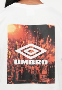 Zadnja stranica bele majice s pomarančno-črno grafiko gorečih industrijskih zgradb in belim logotipom Umbro v središču zasnove.