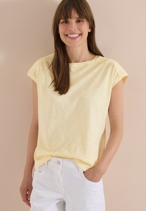 Jeune femme souriante portant un haut à manches courtes jaune clair et un pantalon blanc, une main dans la poche, sur fond beige.