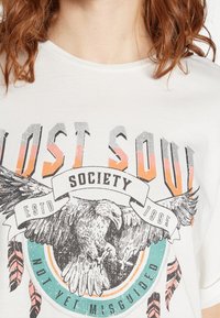 Bílý tričko s grafickým potiskem orla, peří a textu. Design obsahuje šedou, oranžovou a tyrkysovou barvu s vintage vzhledem.