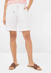Pantaloni corti in lino bianco con vita elasticizzata, chiusura con cordoncino e tasche laterali. Indossati con sandali marroni.
