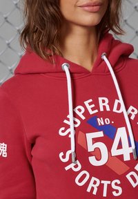 Sudadera roja con capucha y cordones blancos, que presenta un diseño gráfico de "SUPERDRY" y "54" en acentos blancos y azules.