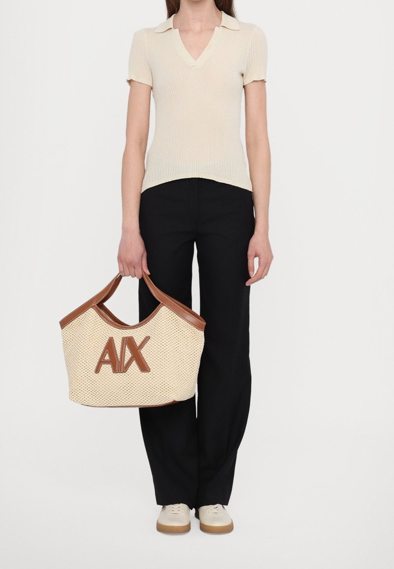 Femme portant un haut côtelé beige à manches courtes, pantalon noir, chaussures claires, tenant un sac fourre-tout tissé beige avec bordure marron et grand logo "A|X".
