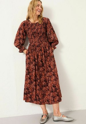 Eine Frau trägt ein langes, gemustertes Kleid in Rostrot und Schwarz mit puffigen Ärmeln, die Hände in den Taschen und silberne Mary Jane-Schuhe, während sie gegen eine weiße Wand steht.