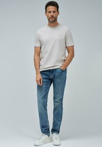 Hombre de pie contra un fondo sencillo, vistiendo una camisa de manga corta beige, vaqueros azul y zapatillas blancas, con una mano en el bolsillo y una expresión neutra.