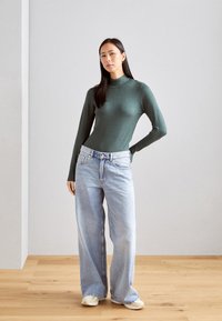 Zelena pletiva turtleneck pulover v kombinaciji s svetlo modrimi širokimi hlačami; ima stranske gumbe in sproščen kroj. Postavljen na nevtralni ozadju.
