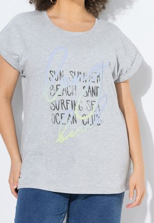 Woman wearing light gray t-shirt with rolled sleeves and pastel text "beach" over black beach-themed words, paired with blue pants.