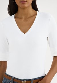 Balts V veida kakla tops ar īsām piedurknēm, izgatavots no ribotiem audumiem. Saskaņots ar brūnu jostu un džinsiem. Vienkāršs dizains ar pieguļošu siluetu.