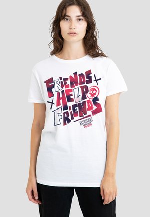 Young woman wearing white T-shirt with red and black text "Friends Help Friends" and Stranger Things logo, standing against a plain gray background.