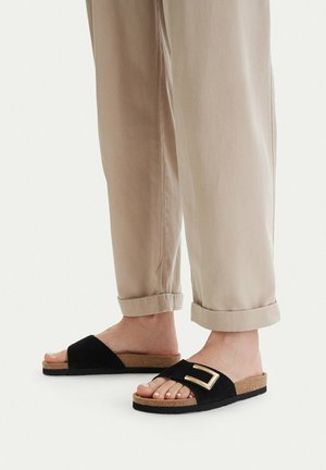 Piedi con pantaloni beige e sandali neri aperti con fibbia dorata, in piedi su una superficie bianca.