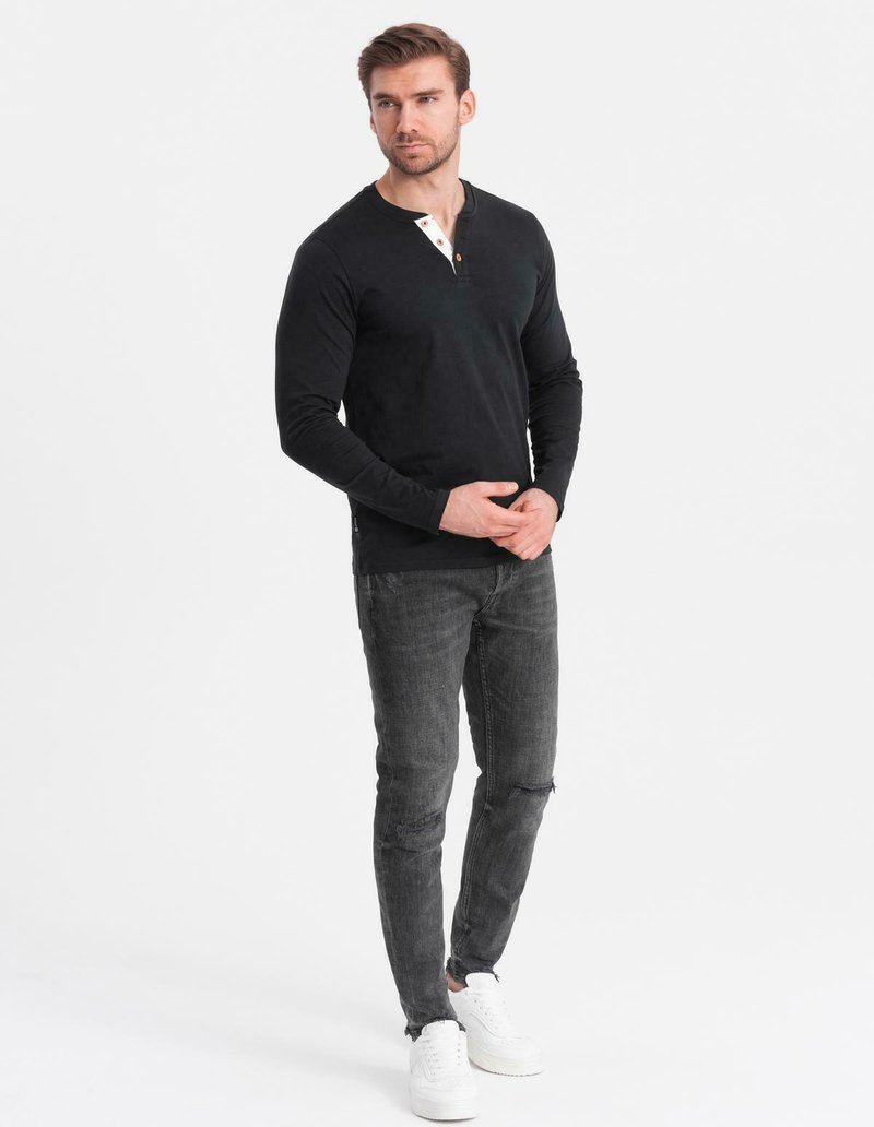 Man med svart långärmad henley skjorta, slitna mörkgrå jeans och vita sneakers står med händerna knäppta och ser något åt höger.