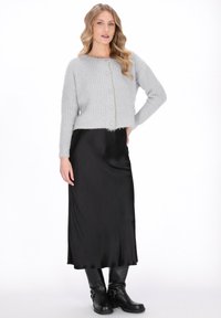 Kobieta o falujących blond włosach, ubrana w jasnoszary sweter z guzikami, czarną satynową spódnicę midi oraz czarne skórzane botki za kostkę.