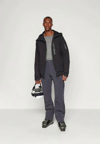 Μαύρο μπουφάν με μόνωση και κουκούλα, γκρι θερμικό πουκάμισο, γκρι παντελόνια χιονιού, μαύρες μπότες σκι, κρατώντας ένα άσπρο και μαύρο κράνος.