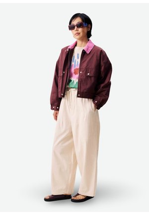Femme portant une veste marron surdimensionnée avec col rose, t-shirt graphique, pantalon large beige, lunettes de soleil surdimensionnées et sandales noires, debout.