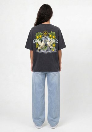 Vrouw draagt een zwarte oversized T-shirt met citroentakken, twee witte duiven en de tekst "Black Bananas", gecombineerd met lichtblauwe wijde jeans.