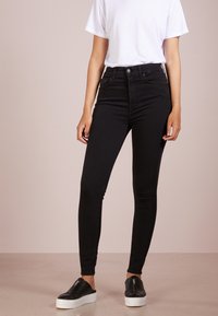 Černé skinny džíny vyrobené z denimu, s vysokým pasem, úzkým střihem a minimálním kovovým doplňkem. Spárované s černými slip-on botami a bílým tričkem.