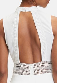 Vit ärmlös klänning med hög bakhalsskärning, som har intrikata spetsdetaljer längs den övre delen av ryggen och en detaljerad spetsaccent vid midjan.