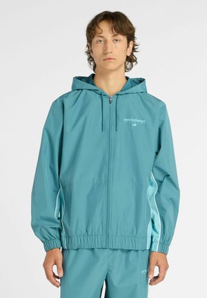 Junger Mann, der eine türkisfarbene New Balance Kapuzenjacke mit Reißverschluss und passende Hose trägt, vor einem schlichten hellen Hintergrund steht.