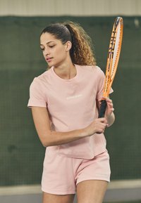 Svetlo roza športna majica in kratke hlače z logotipom, v roki pa drsni teniški lopar z teksturo. V ozadju je vidno teniško igrišče.