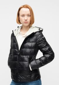 Νεαρό άτομο με κοντά κόκκινα μαλλιά, φοράει μαύρο επενδυμένο μπουφάν με κουκούλα πάνω από λευκό πουκάμισο και μπλε τζιν, με τα χέρια στις τσέπες του μπουφάν, ουδέτερο φόντο.