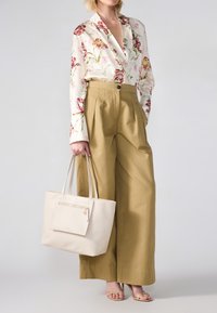 Blouse à manches longues à motifs floraux avec des couleurs vives, pantalons beiges taille haute et sac fourre-tout léger texturé avec zip. Fond clair.