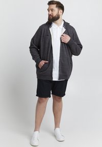 Grå zip-up hettegenser med hvit indre fôr, to frontlommer, brukt over en hvit polo skjorte, parret med svarte shorts og hvite joggesko.