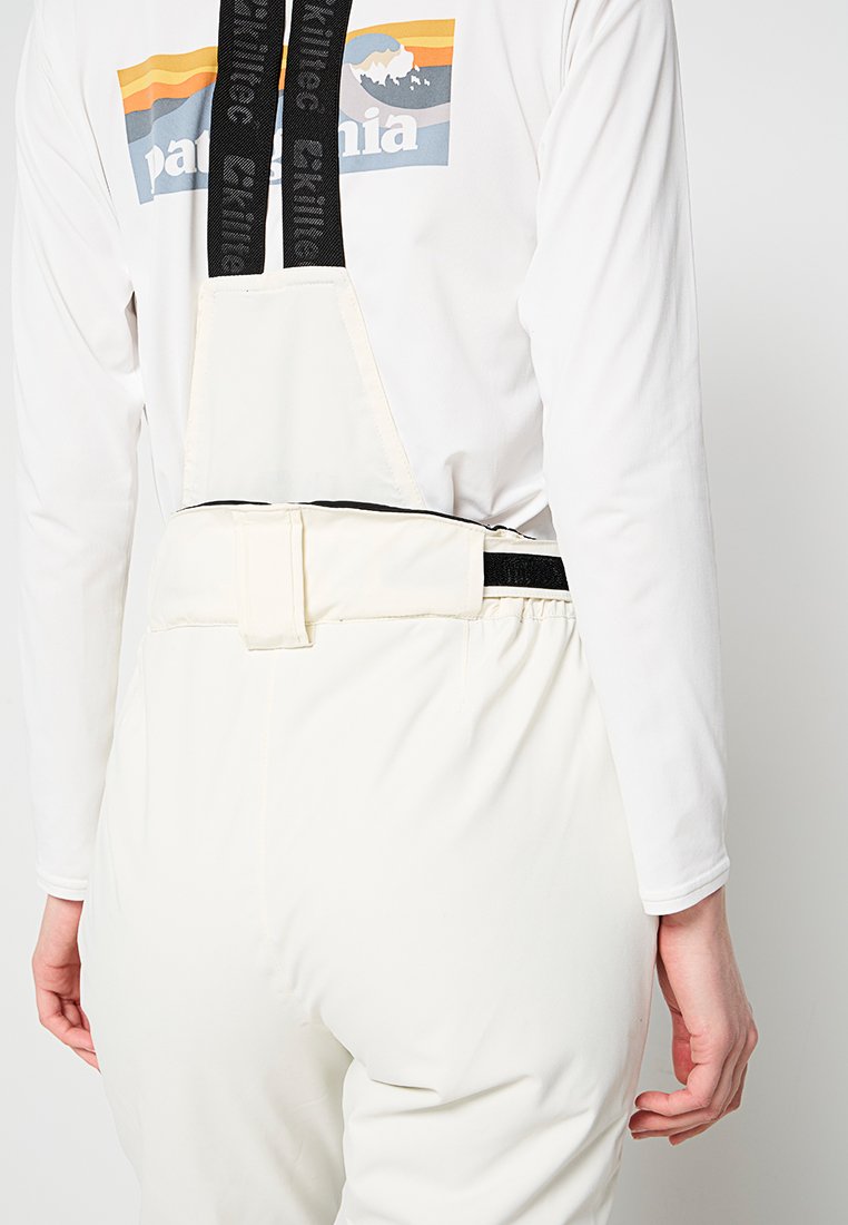 Witte overall met een zwarte verstelbare riem en klittenbandsluiting, gecombineerd met een wit shirt met lange mouwen en een kleurrijk Patagonia-logo.