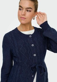 Mornarska pletena cardigan jakna s teksturiranim dijamantnim uzorkom, pet gumba i vezanim strukom. Izrađena od mekanog konca s rebrastim manžetama.