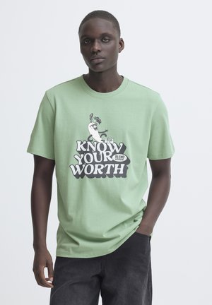 Giovane uomo che indossa una maglietta verde chiaro con un cartone animato di una verdura e il testo "Know Your Worth", in piedi con una mano in tasca.